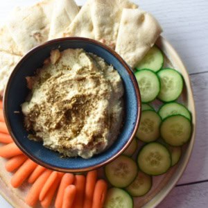 Artichoke hummus in a blue bowl surrounded by cucumber, carrots, and pita bread on a white surface