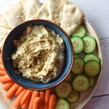 Artichoke hummus in a blue bowl surrounded by cucumber, carrots, and pita bread on a white surface