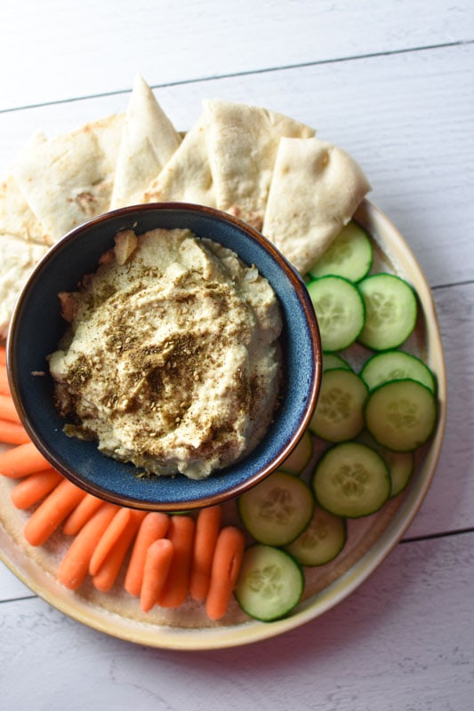 Artichoke hummus in a blue bowl surrounded by cucumber, carrots, and pita bread on a white surface