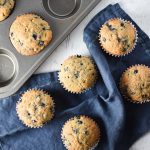 Blueberry muffins on a table next to a muffin pan and blue towel
