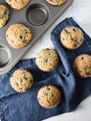 Blueberry muffins on a table next to a muffin pan and blue towel
