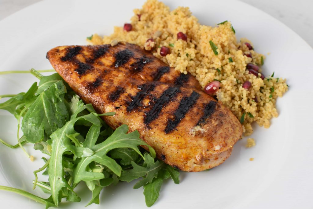 Grilled chicken next to couscous with pomegranate seeds and green arugula on a white plate