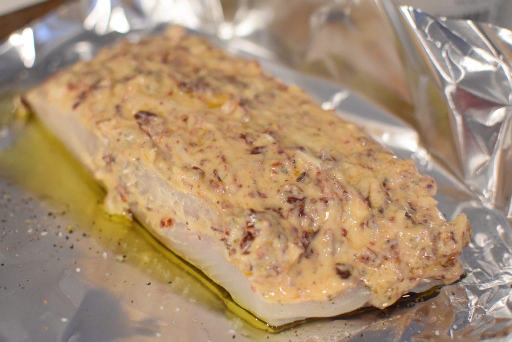 A prepped piece of halibut topped with a mayonnaise mixture of sundried tomatoes and spices on a baking sheet.