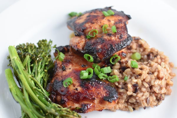 Honey garlic chicken thighs with rice and broccolini, topped with green onions, on a white plate