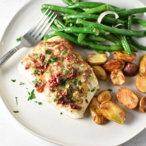 A filet of mediterranean baked halibut on a white plate with potatoes and green beans.