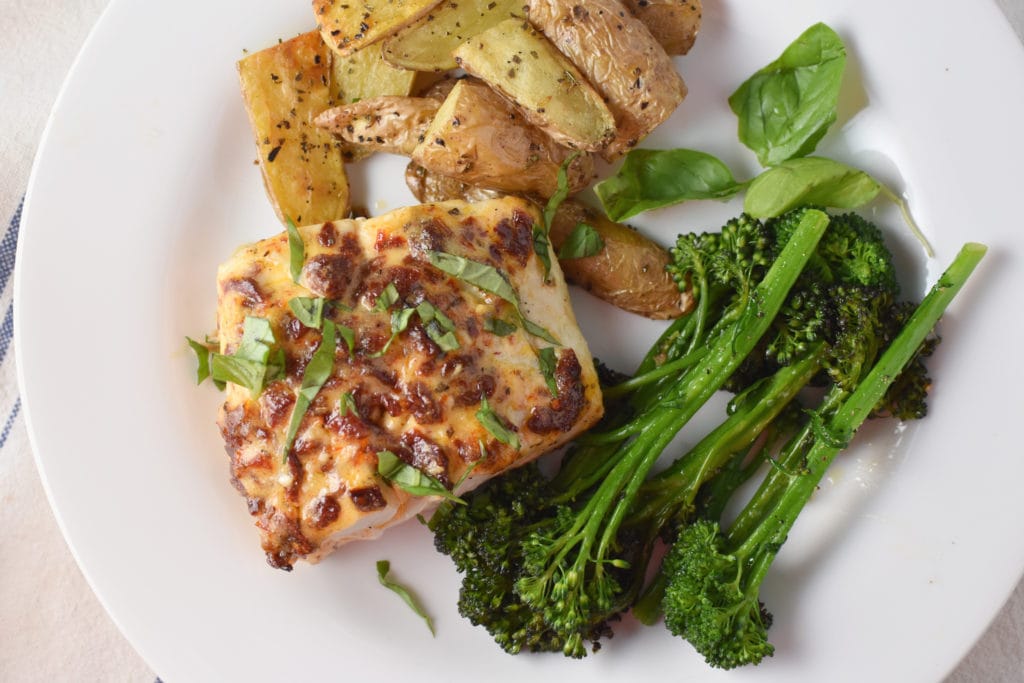 A piece of halibut topped with tomatoes, basil, and olive oil next to roasted potatoes and broccolini