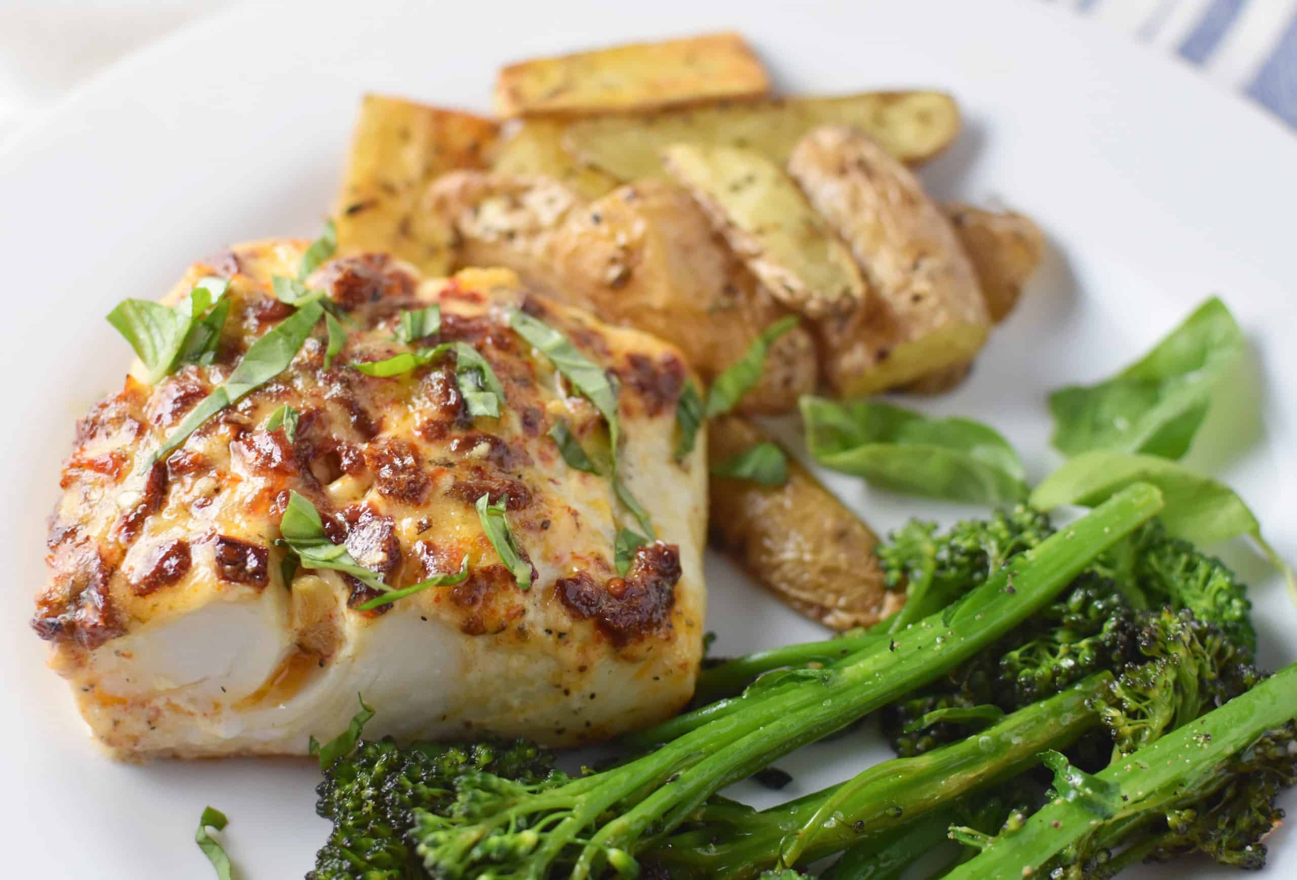 Mediterranean baked halibut on a white plate with broccoli and potatoes with a dish towel in the back