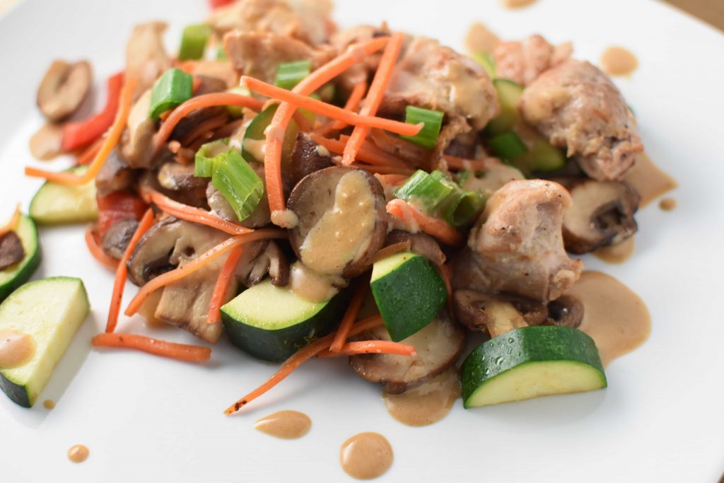 A white plate with chicken stir fry covered in a sesame sauce and topped with carrots. 