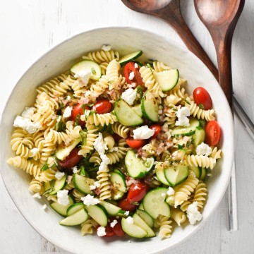 A white bowl of gluten free pasta salad with tomatoes and zucchini next to salad tongs