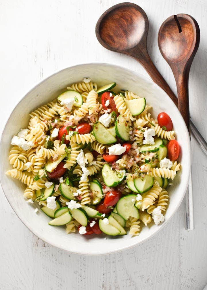 A white bowl of gluten free pasta salad with tomatoes and zucchini next to salad tongs