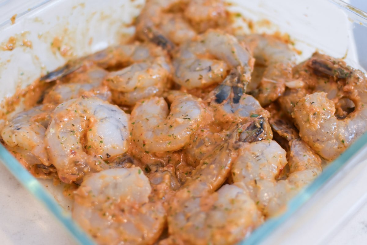 Raw shrimp being marinated in a tomato based sauce in a glass container