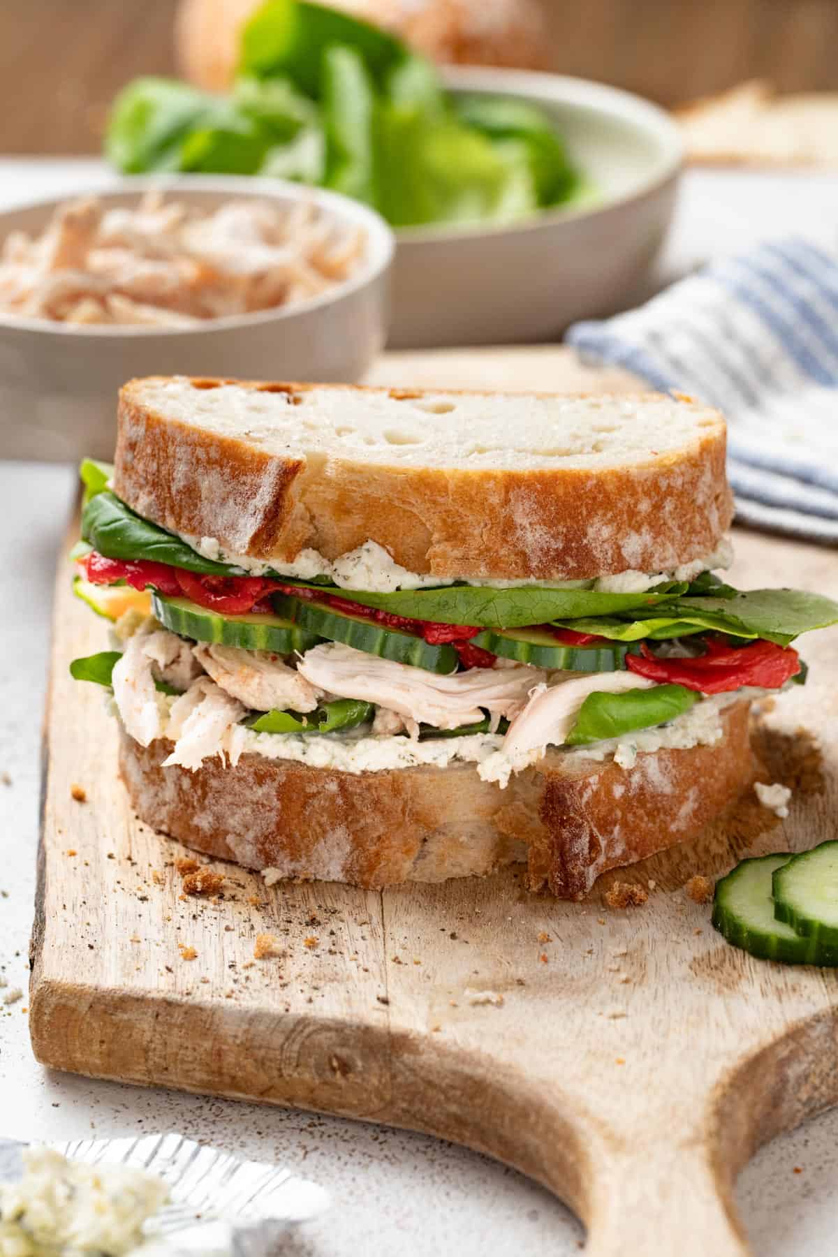 A layered chicken sandwich on top of a cutting board next to a bowl of rotisserie chicken and lettuce.