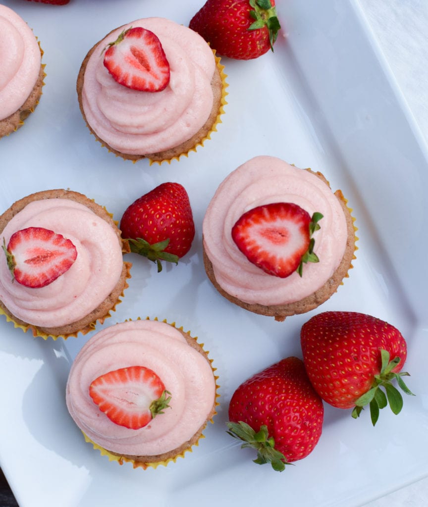 Cupcakes with strawberry frosting and strawberry halves on top on a white plate.