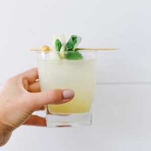 A hand holding a pear mojito mocktail up against a white wall.