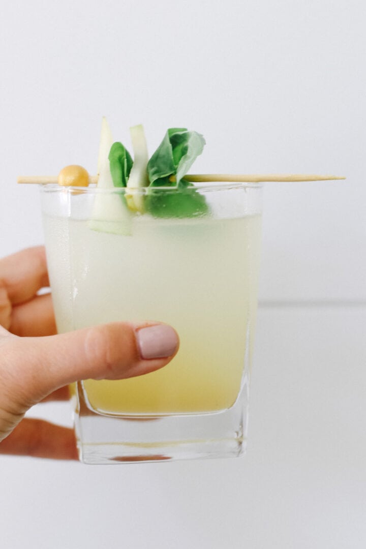 A hand holding a pear mojito mocktail up against a white wall.