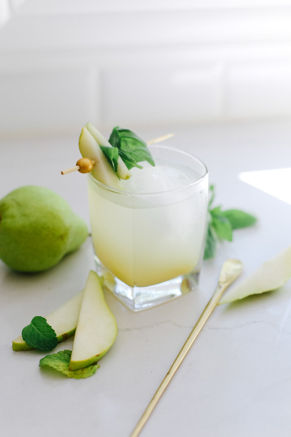 A pear mocktail next to a gold cocktail stirrer, sliced pears, mint leaves, and a cocktail glass..