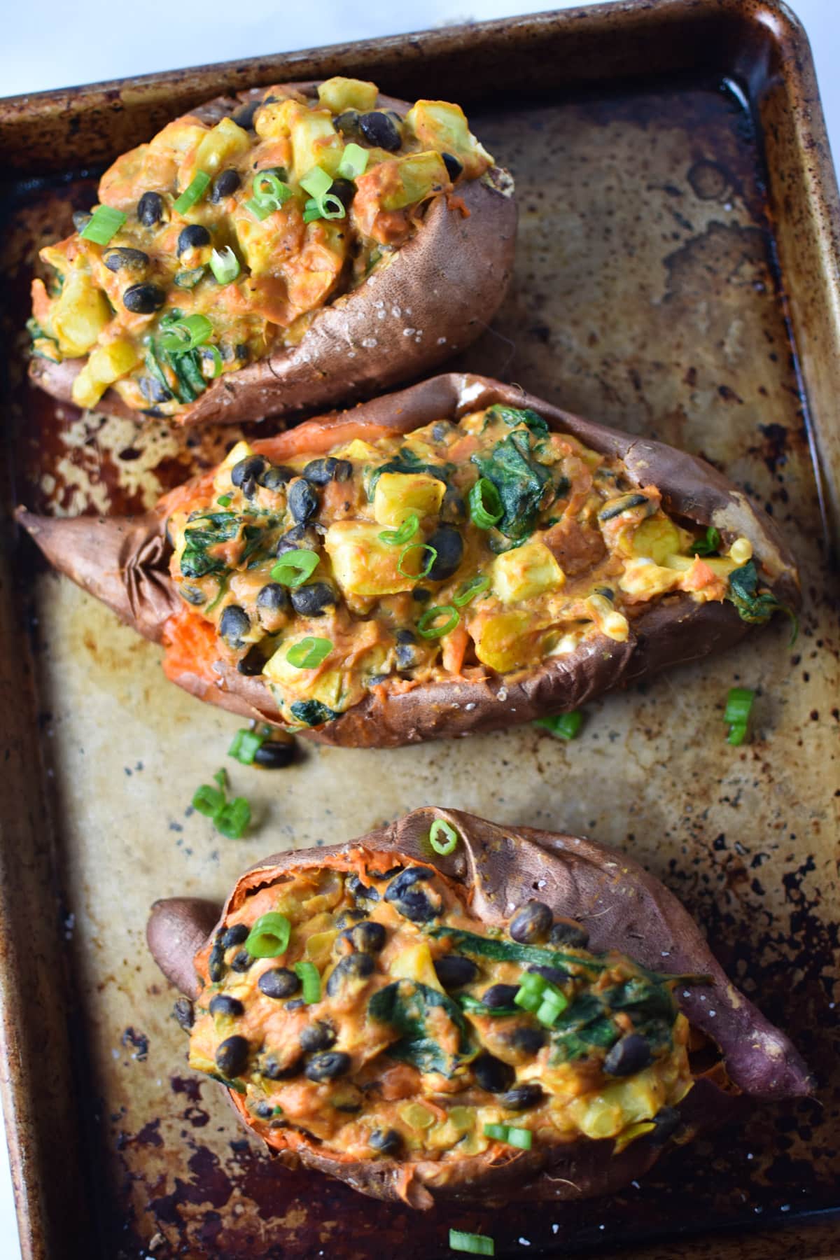 A sheet pan with three stuffed sweet potatoes topped with black beans, squash and green onion.