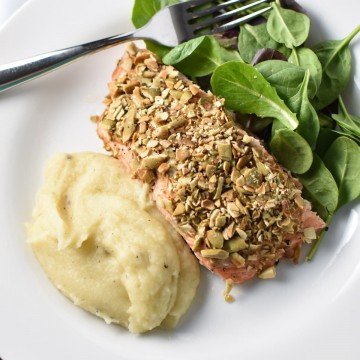 Pepita crusted salmon on a white plate with a fork and mashed potatoes with lettuce