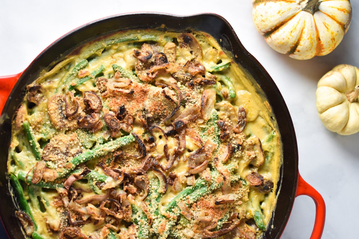 A green bean casserole with shallots in a cast iron pan next to two pumpkins.