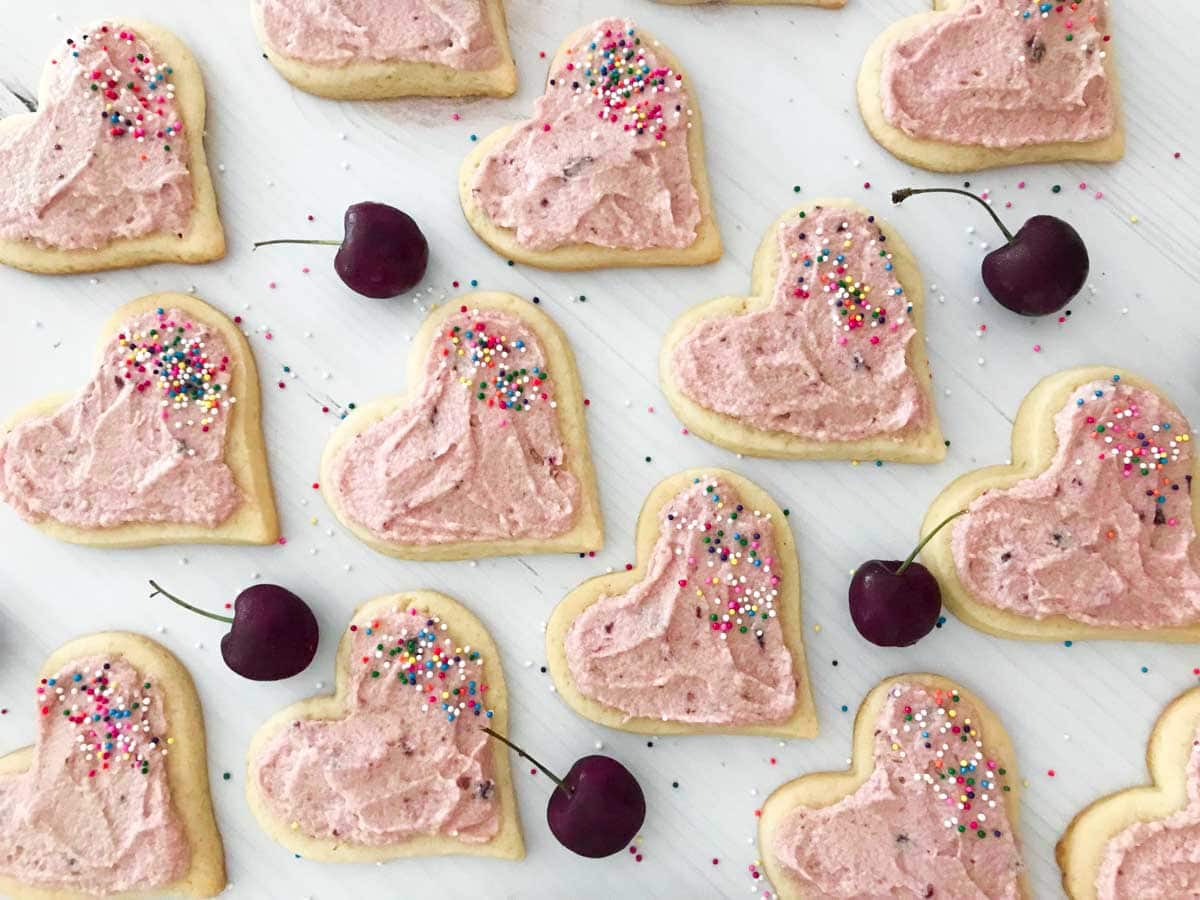Heart shaped sugar cookies with cherry frosting and sprinkles on a white table with fresh cherries