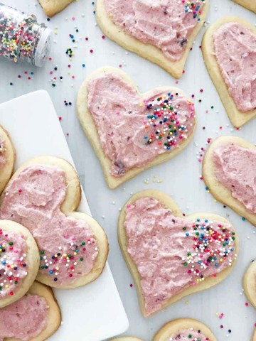 Heart shaped sugar cookies with pink cherry frosting and sprinkles on a plate with sprinkles scattered on top