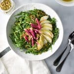 Pear arugula salad on a white table with a napkin, serving spoons, and dressing