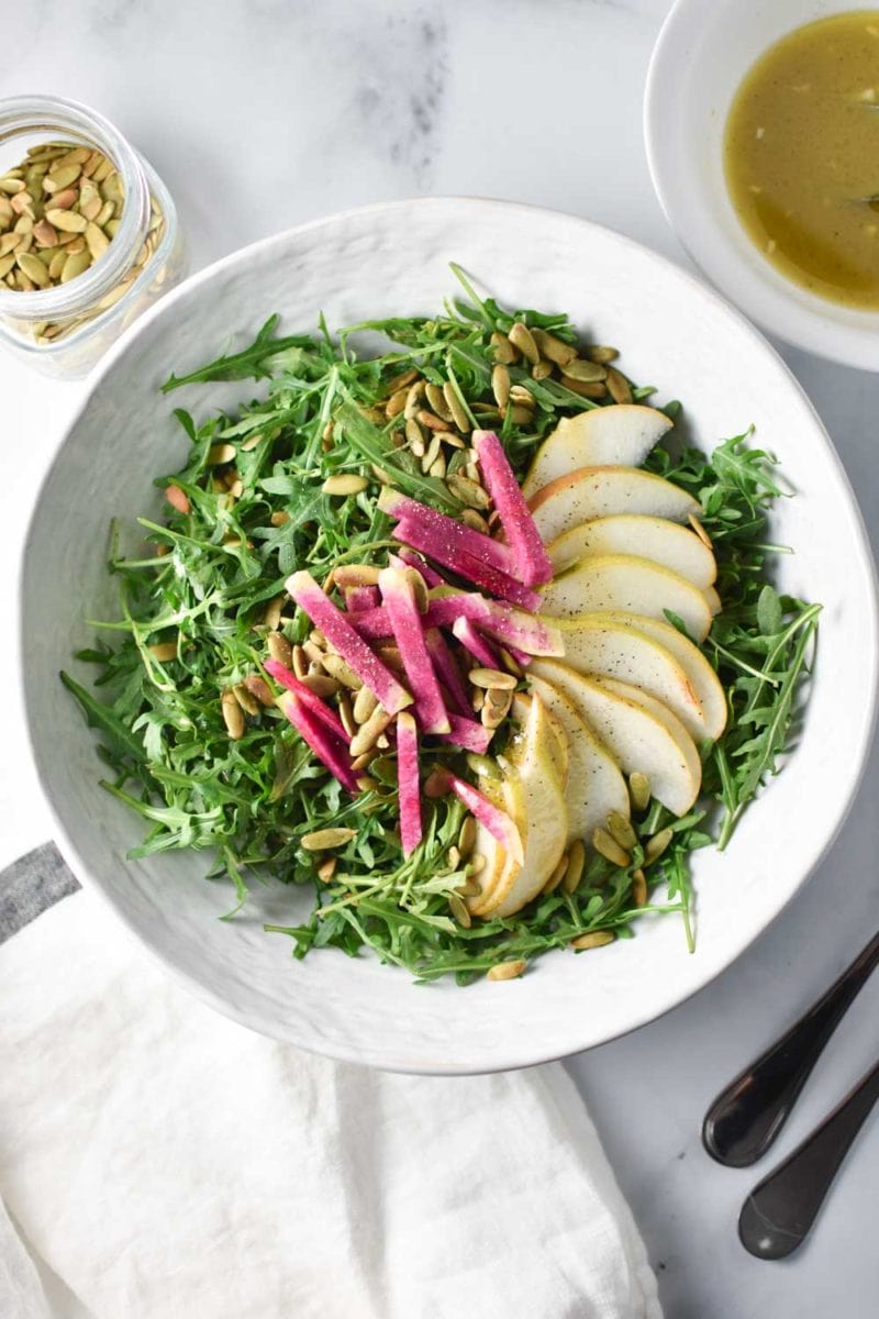 Arugula salad with pears and radish in a white bowl surrounded by pepitas and dressing.