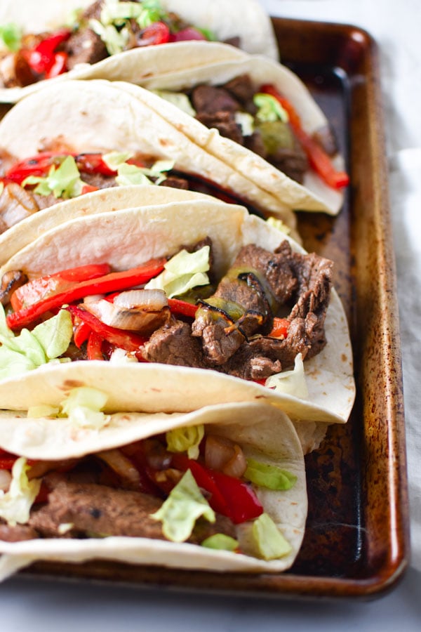 A line of packed steak fajitas piled high with flank steak meat, shallots, and peppers on a pan