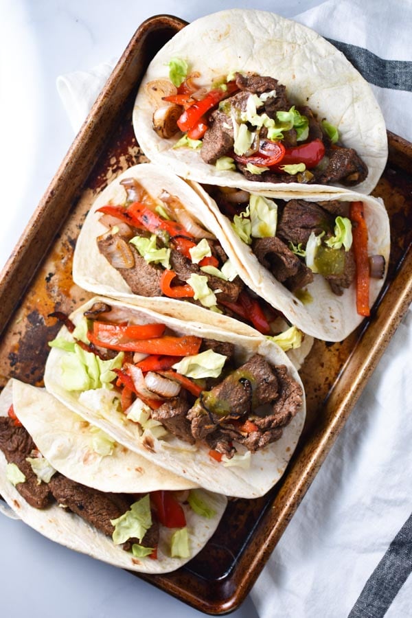 Healthy steak fajitas topped with shallots and red pepper in tortillas on a sheet pan