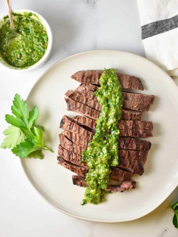 Sliced flank steak on a plate with chimichurri sauce on top and parsley