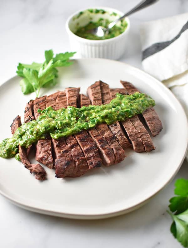 Sliced grilled steak with herb chimichurri on top on a marble table with a bowel of chimichurri in the background