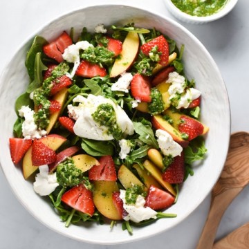 Burrata, peach, and strawberries in a salad bowl with basil vinaigrette in a white cup and tongs