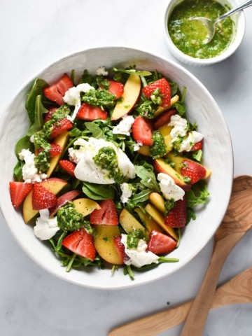 Burrata, peach, and strawberries in a salad bowl with basil vinaigrette in a white cup and tongs