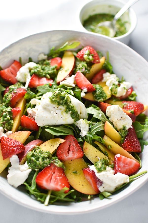 Peaches, strawberries, and burrata on arugula in a white bowl with a basil vinaigrette in the background 