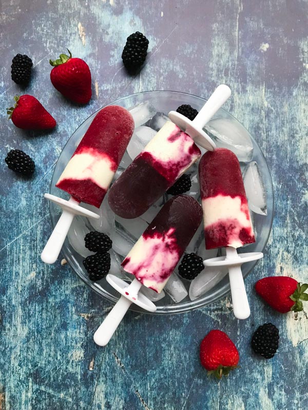 Berry cheesecake popsicles on top of a bowl with ice cubes and wooden background with strawberries and blackberries