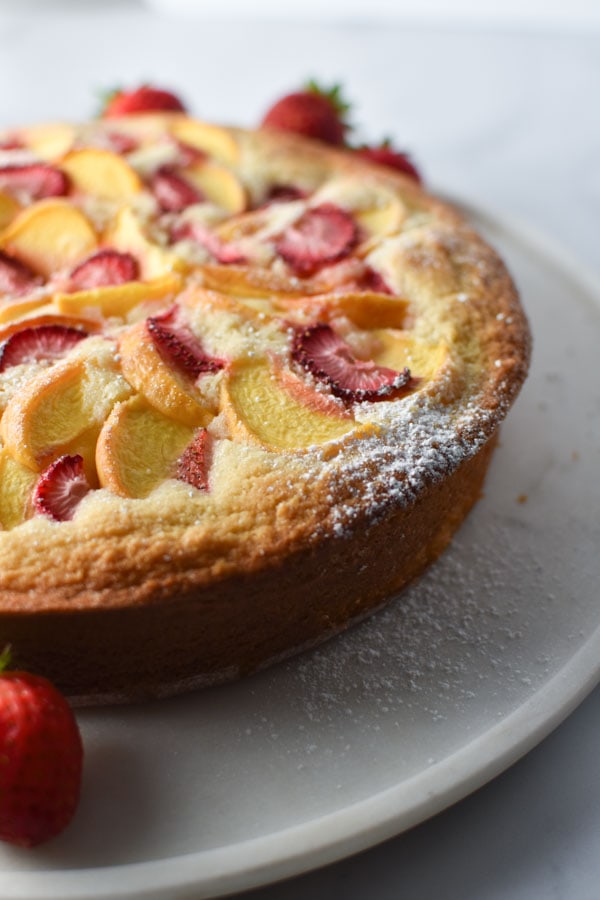 Side view close up of vanilla cake with peaches and strawberries topped with powdered sugar on a white marble plate