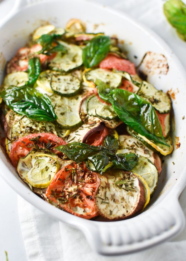 A baked vegetable tian that is topped with fried crispy basil in a white dish