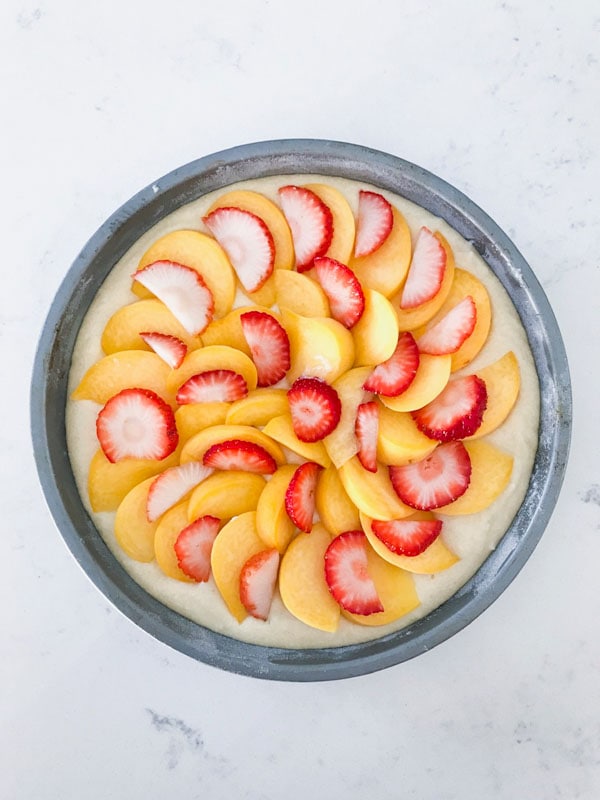 Peaches and strawberries layered on top of a cake in a silver cake pan on a white surface