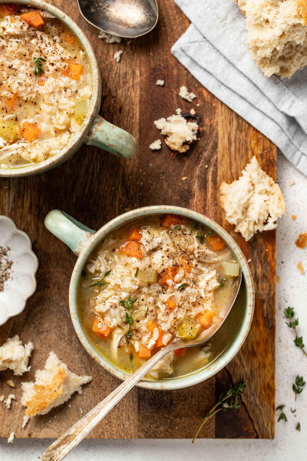 Two bowls of chicken soup with rice on a wood board with crusty bread.