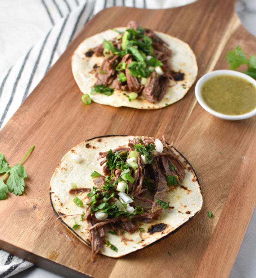 Shredded beef tacos on a wood board with cilantro and salsa verde