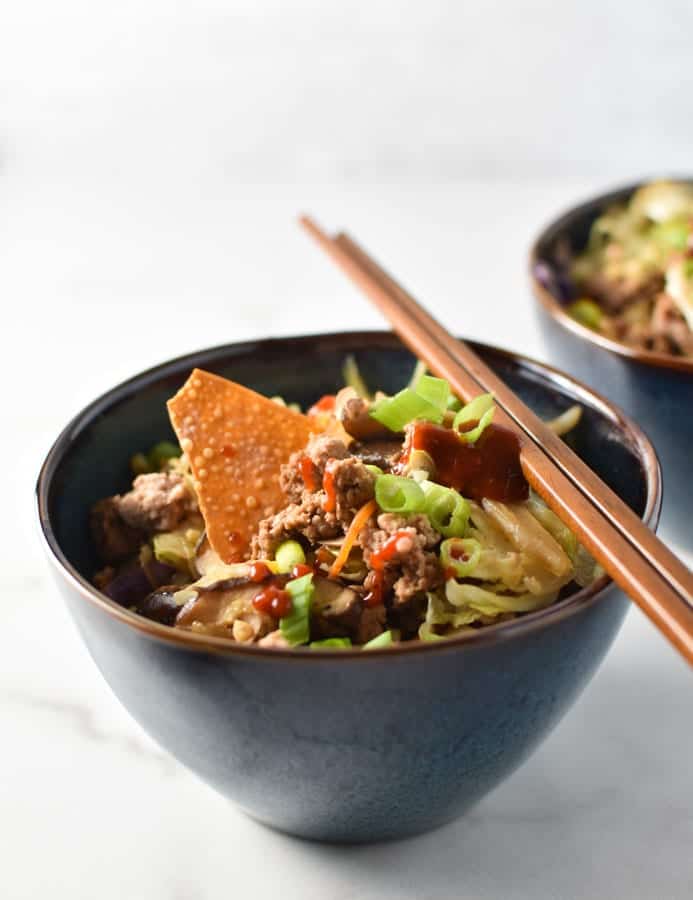 an egg roll bowl with a wonton chip and sriracha on top with chopsticks