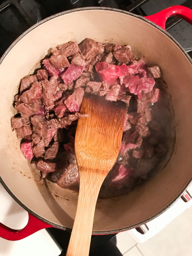 A wood spoon stirring a big pot of meat in a dutch oven.