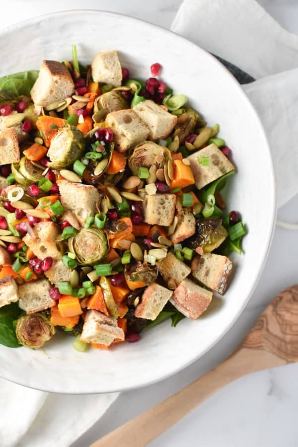 Roasted vegetable salad in a white bowl with wooden serving tongs.