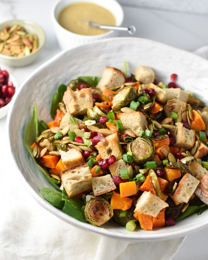 A roasted fall panzanella salad in a white bowl with dijon dressing and pepitas off to the side