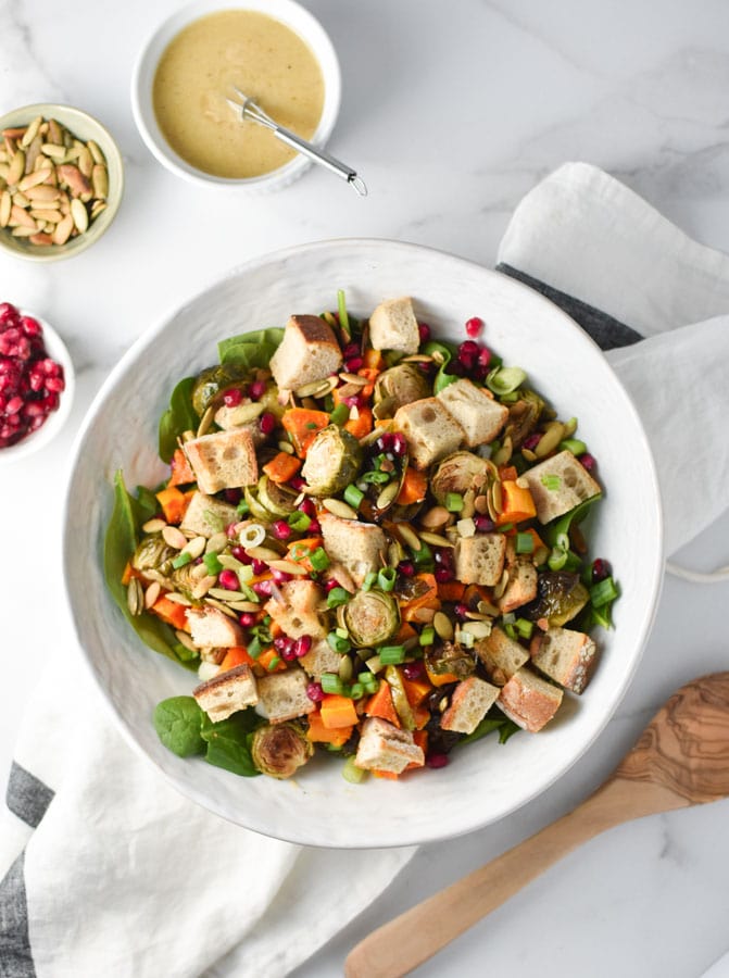 winter roasted vegetable salad with croutons on top in a white bowl with dressing, pepitas, and pomegranate seeds in little bowls