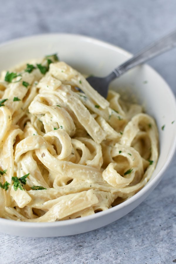 Pasta in a cream sauce twirled around a fork in a white bowl, topped with parsley.