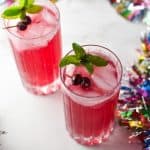 Two cocktail glasses filled with ice and a cranberry mocktail topped with cranberries and mint on a white marble surface