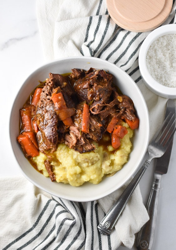 Pot roast, carrots, and mashed potatoes in a bowl on top of a striped towel with a knife and fork to the side