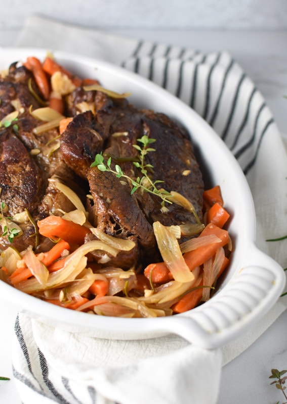 A Whole30 pot roast in a white roasting dish with carrots and shallots