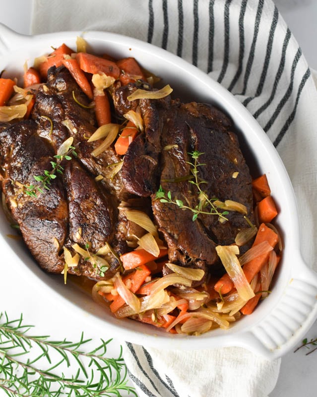 A roasting dish with pot roast, carrots, and shallots, with rosemary sprigs on a white table